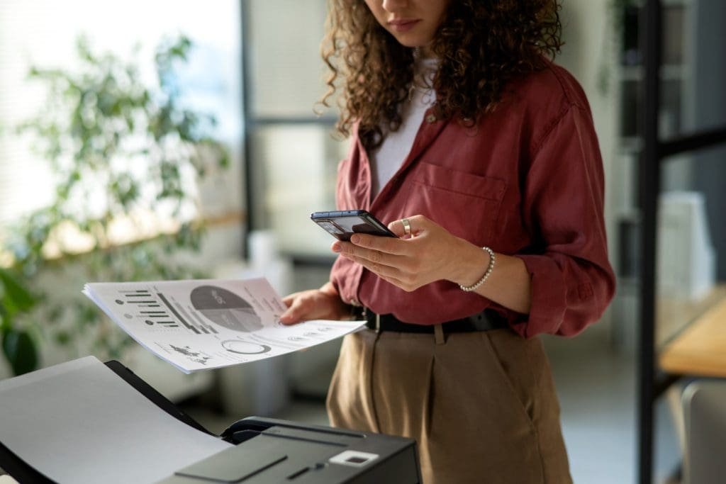 Person using a smartphone while printing a business report in a modern office setting.